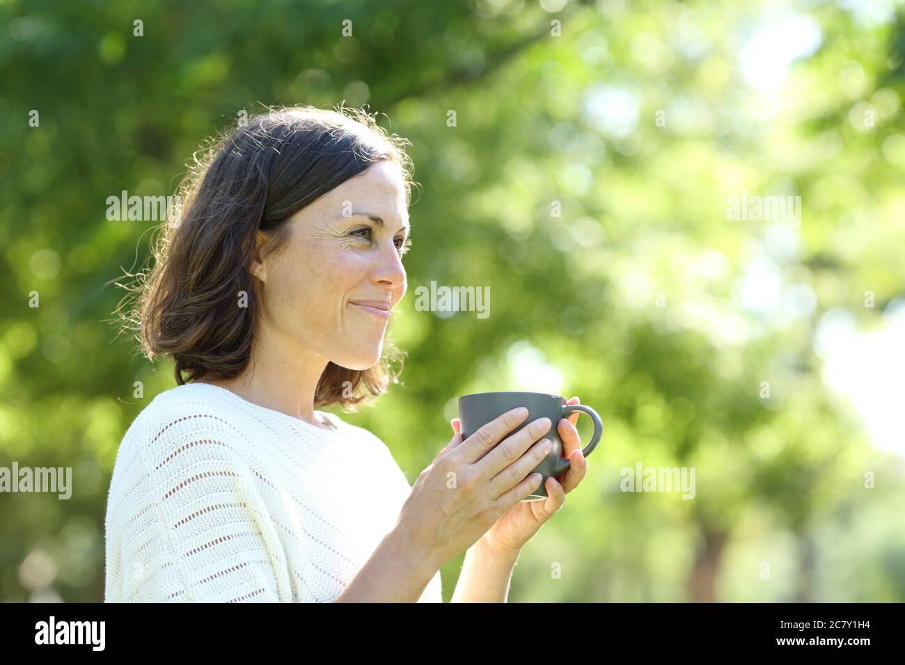 Satisfied adult woman holding a coffee cup standing in the park at summer Stock Photo