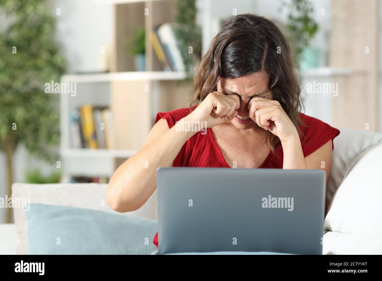 Adult woman with laptop scratching itchy eyes sitting in the sofa in the living room at home Stock Photo