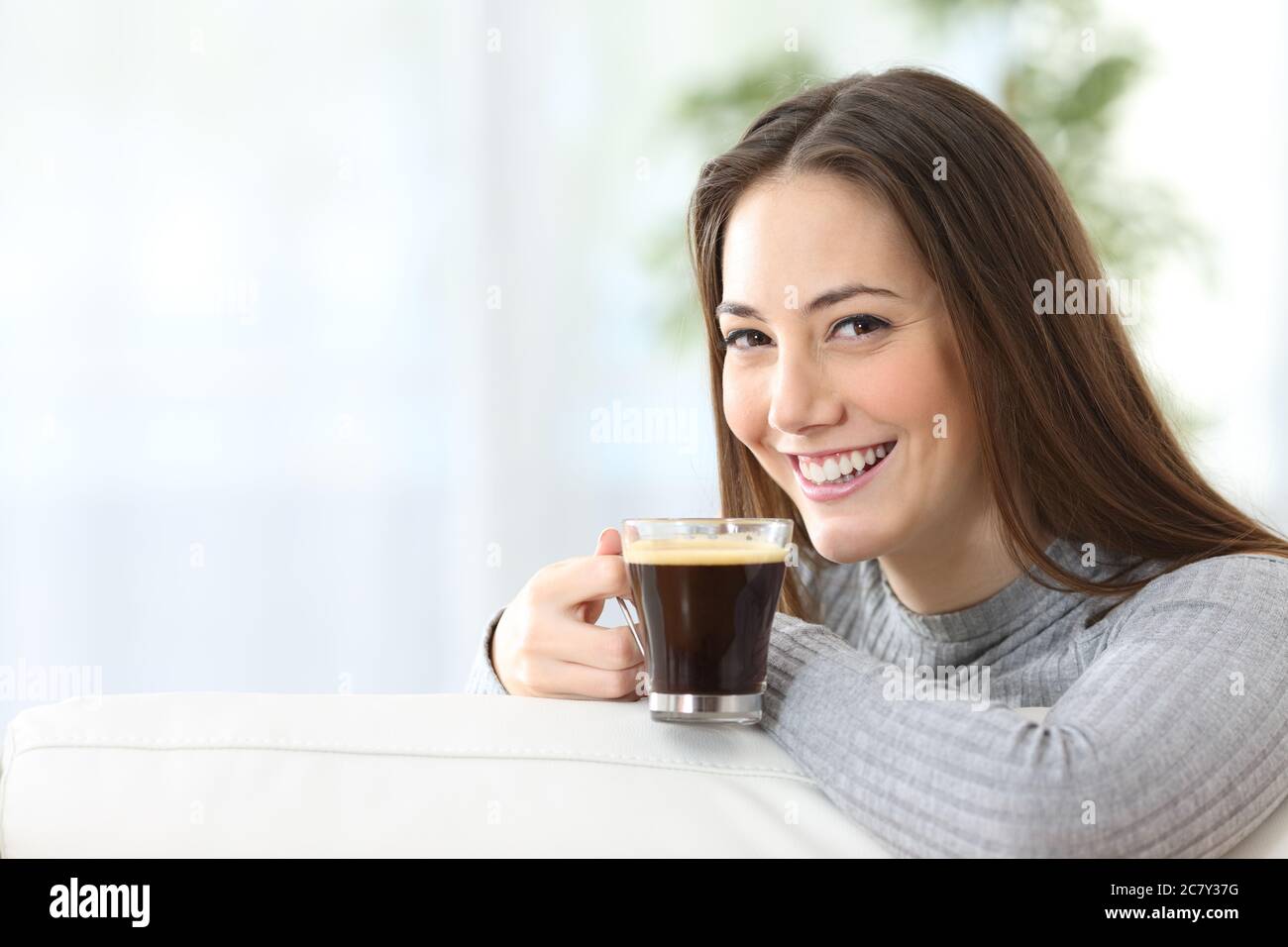 Happy woman with coffee cup looking at camera sitting in the sofa at home Stock Photo