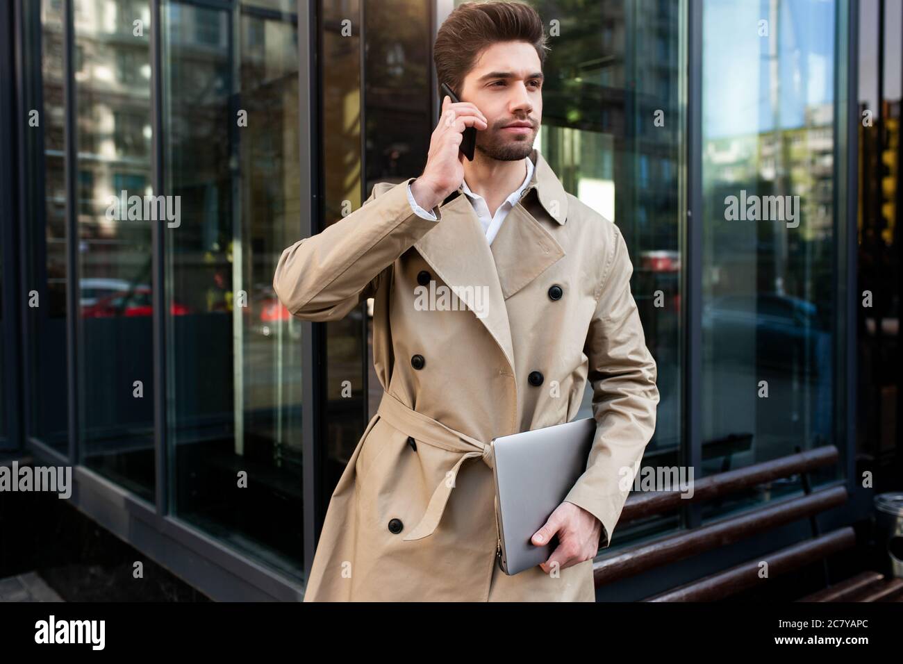 Young casual businessman in trench coat with laptop confidently talking on  cellphone on street Stock Photo - Alamy