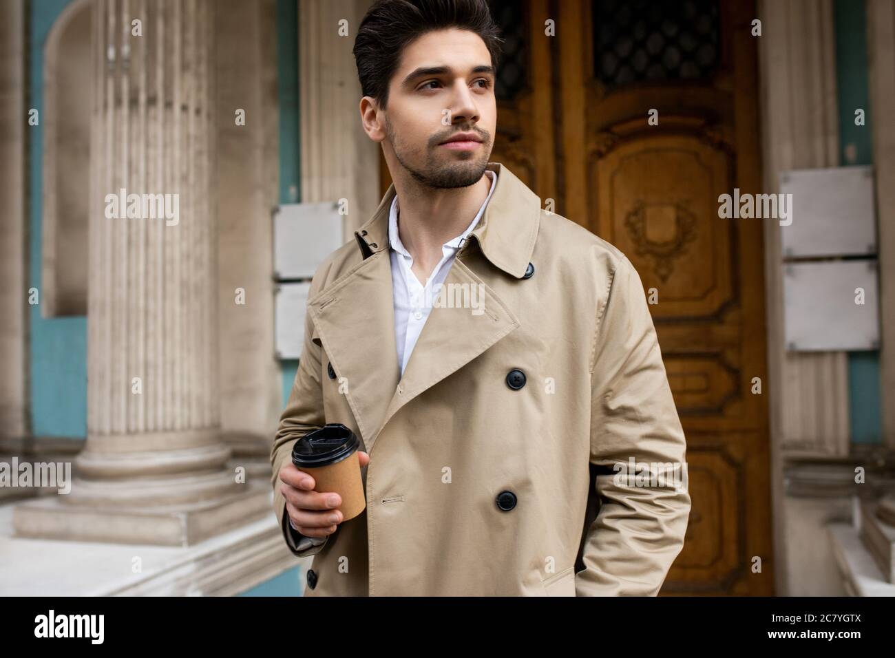 Young attractive man in trench coat with coffe to go dreamily looking away walking through street Stock Photo