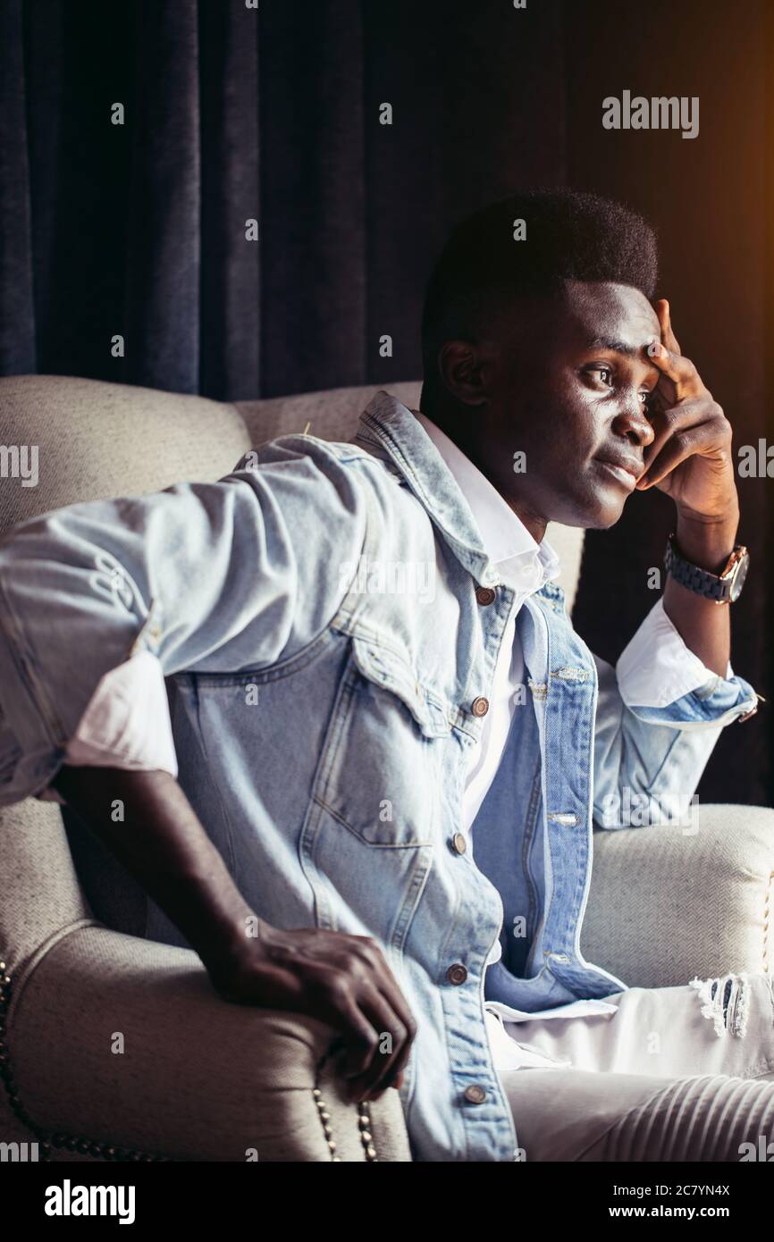 Profile of african young man in light blue denim jacket sitting in armchair, looking pencively Stock Photo