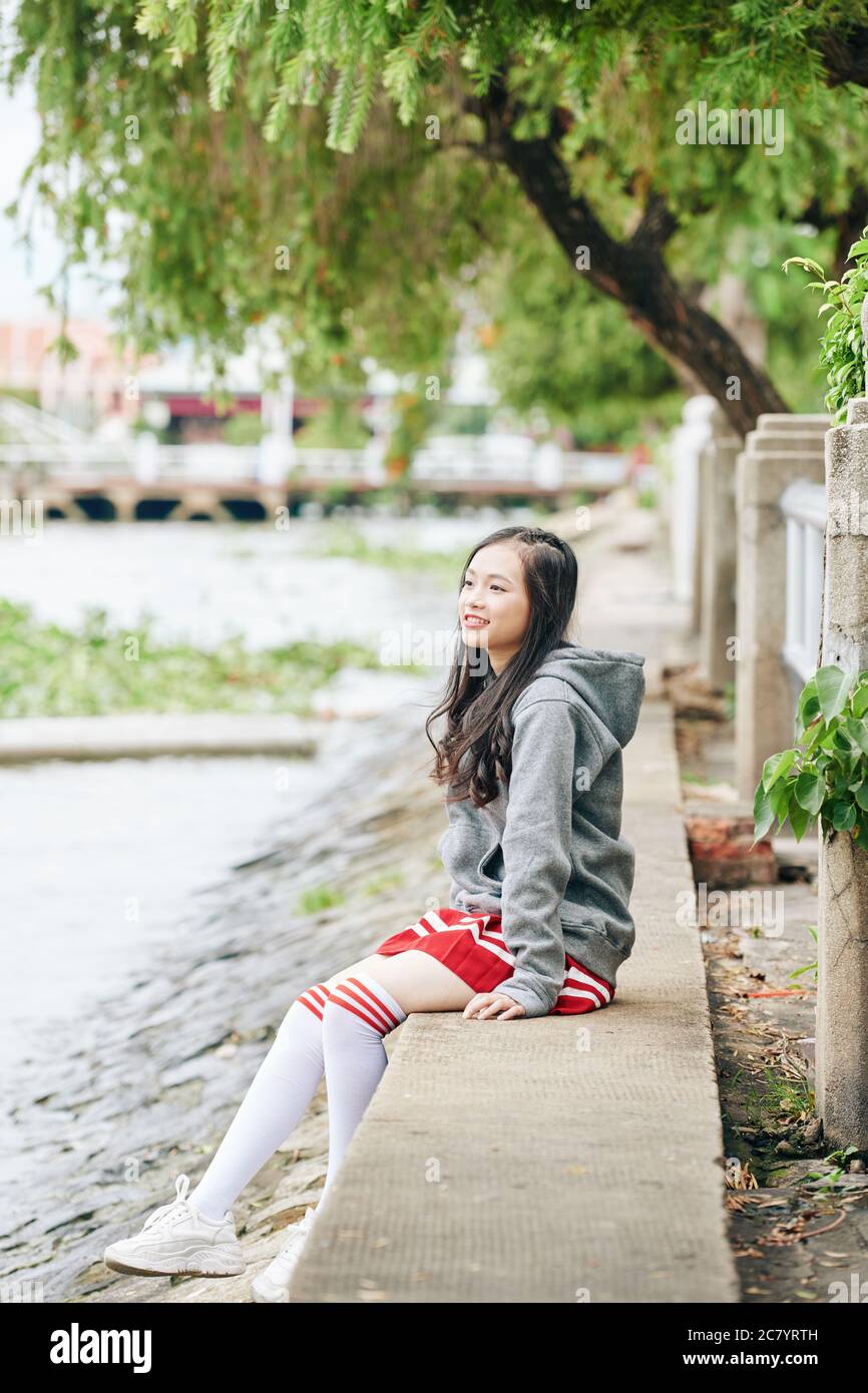 Smiling lovely young Asian woman in skirt, hoodie and thigh high socks  sitting on river bank and looking at water Stock Photo - Alamy