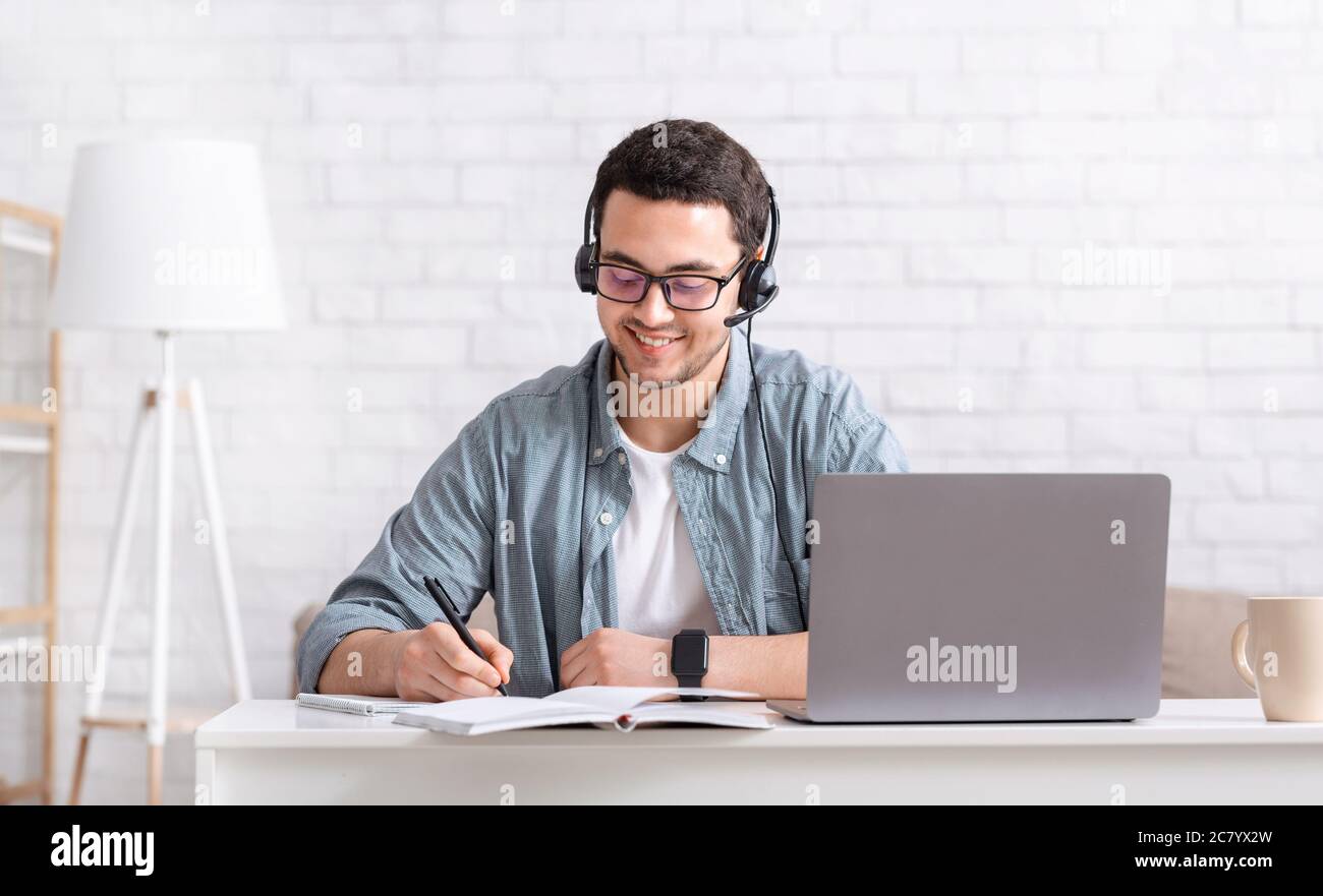 Education remotely and independently. Smiling guy with headphones makes notes in notebook with laptop on desktop Stock Photo