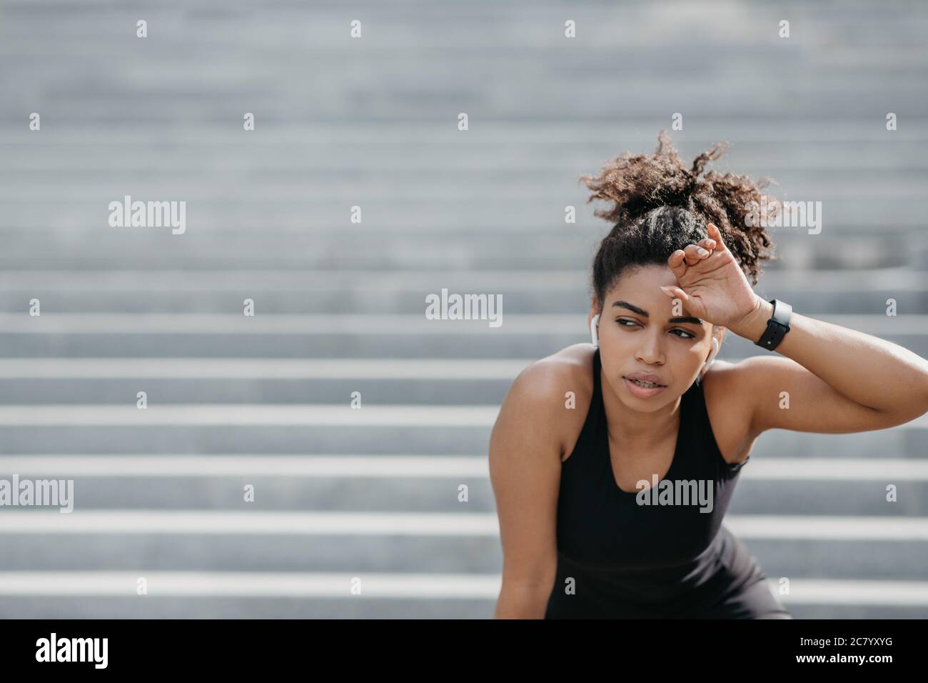 Tired girl in sportswear and smart watches wipes sweat from forehead on stairs background Stock Photo
