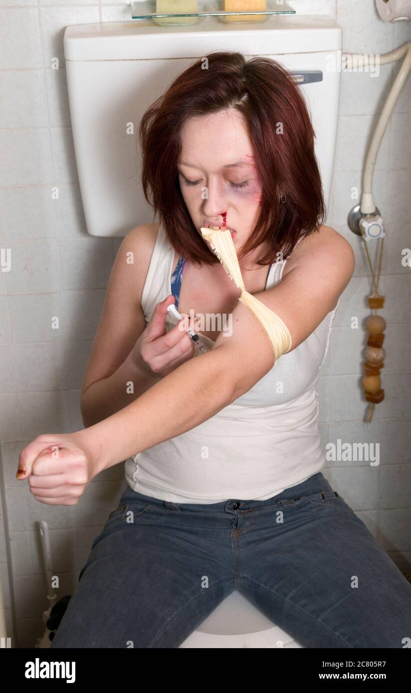 Young woman in her 20s, heroin addict shooting up herself with a syringe, in the arm, in an household toilet Stock Photo
