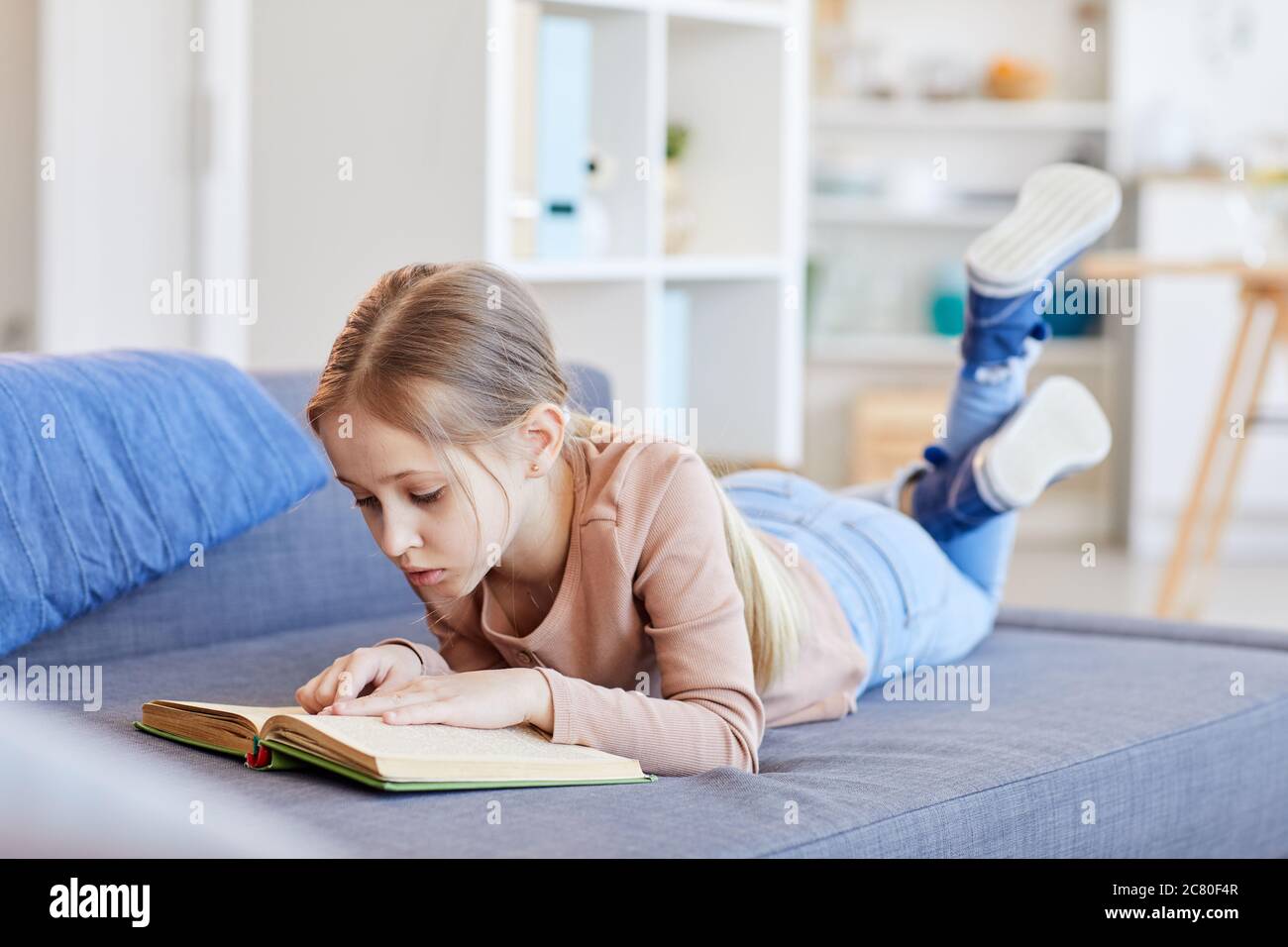 Full length portrait of cute blonde girl reading book or studying while lying on couch in cozy home interior, copy space Stock Photo