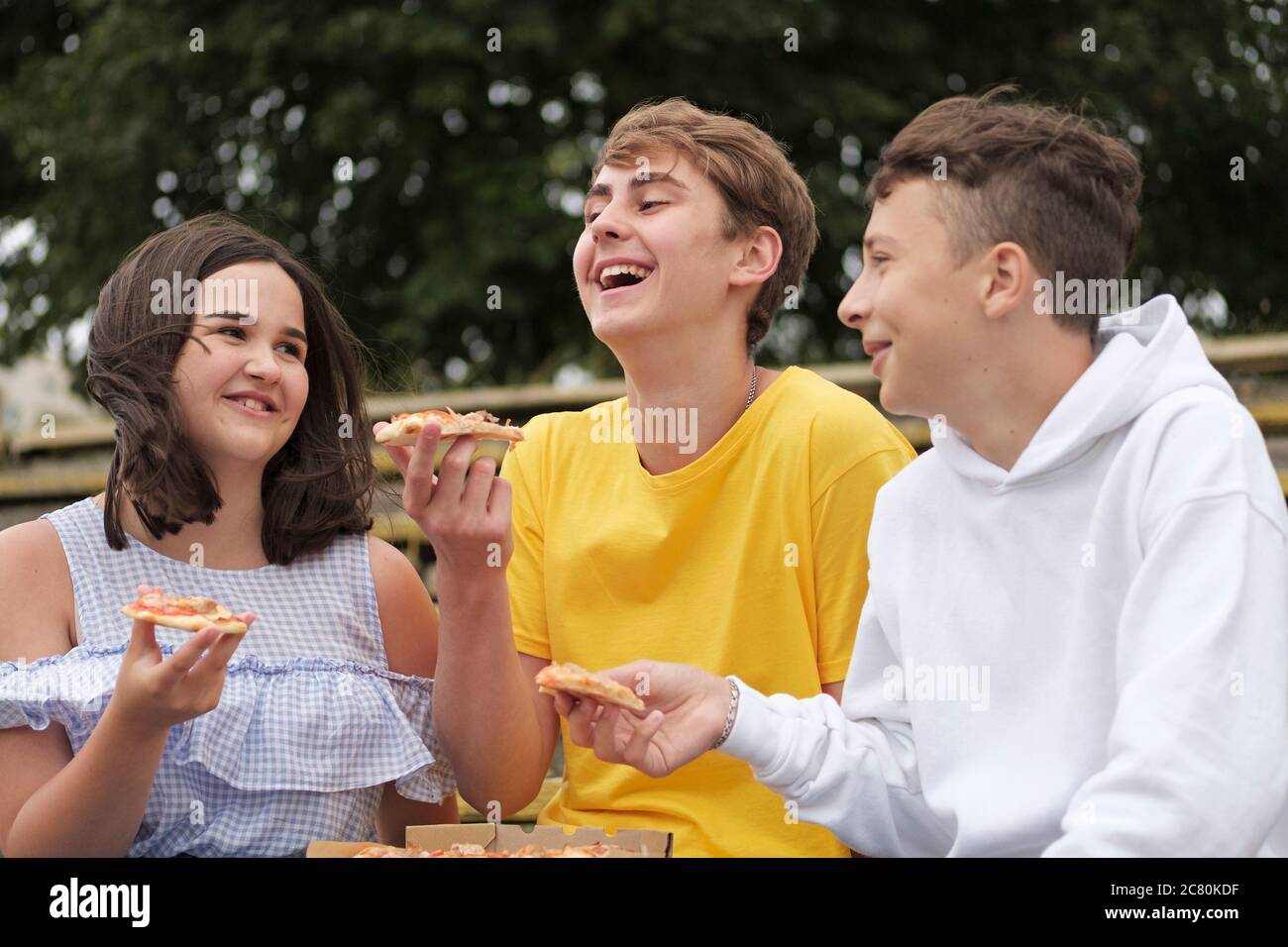 Two teenage boys and a girl sharing snacks outdoors laughing and joking together as they eat in a close up portrait Stock Photo