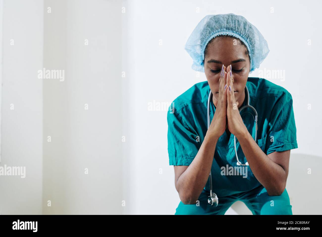 Tired young Black medical nurse praying for sick and suffering people when sitting on the floor in hospital corridor Stock Photo