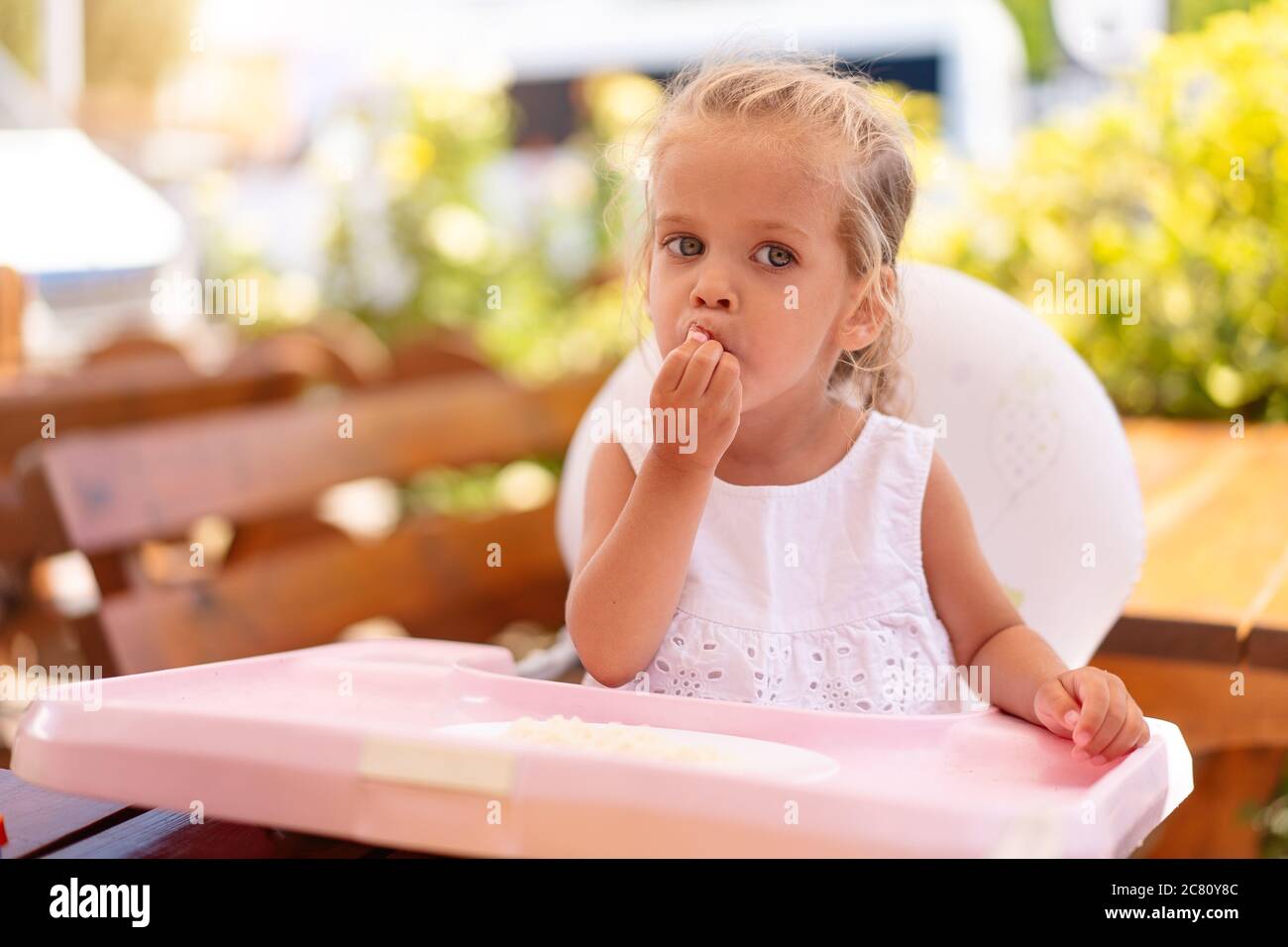 Cute little Caucasian girl eating spaghetti at table sitting in child seat outdoor restaurant. Healthy eating. Funny child eat street food sunny summer day. Stock Photo