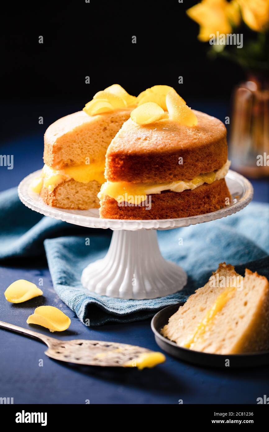 Decadent vegan lemon sponge cake presented on decorative white cake stand, topped with sugared yellow rose petals.  Slice of cake on plate Stock Photo