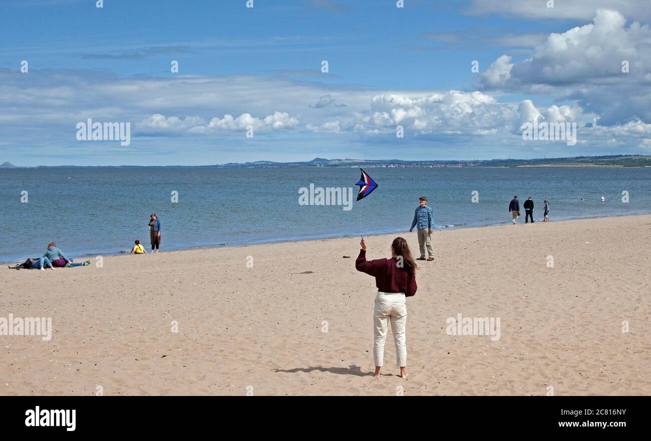 Portobello, Edinburgh, Scotland, UK. 20 July 2020. This young woman having fun flying her kite over a very quiet beach for the middle of July. Weather sun and cumulus clouds with windy conditions. Wind  West 23 km/h gusts 37 km/h temperature of 17 degrees. Stock Photo