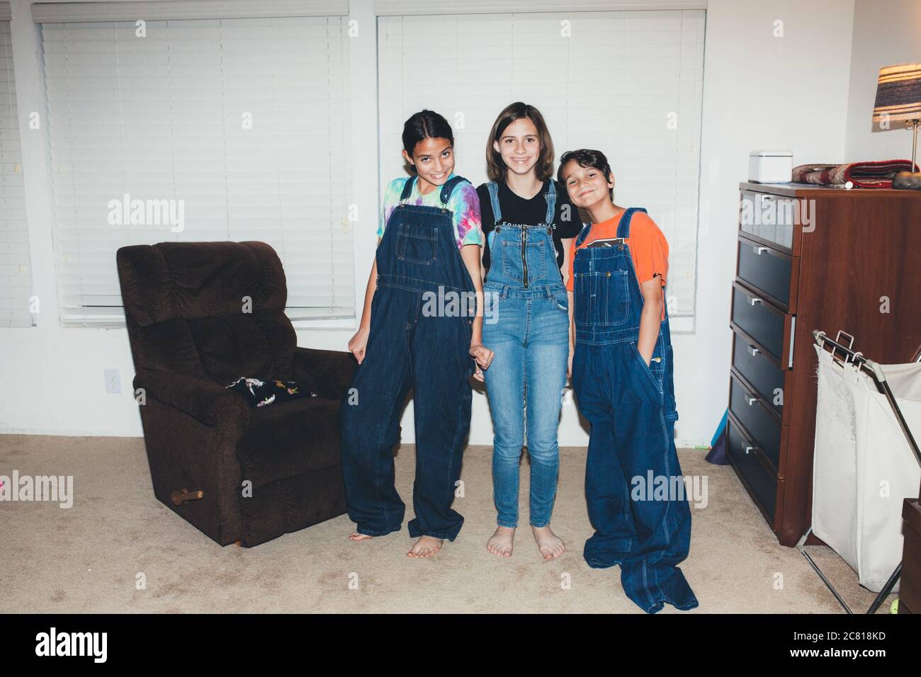 Three siblings smile while all wearing overalls and two oversized Stock Photo