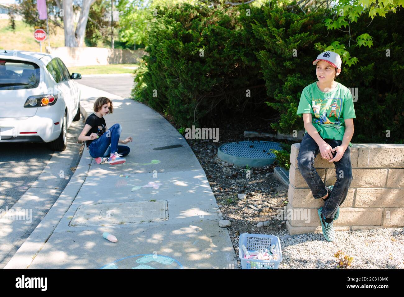 Boy sits on a wall while his sister does chalk art on the sidewalk Stock Photo