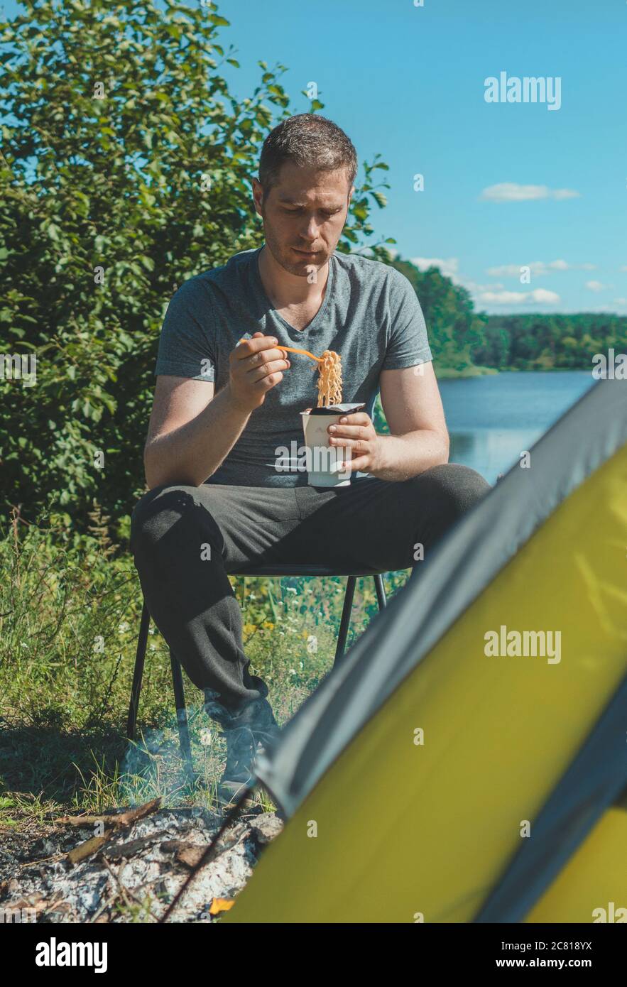 Man is eating instant noodles next to tent by the lake. Stock Photo