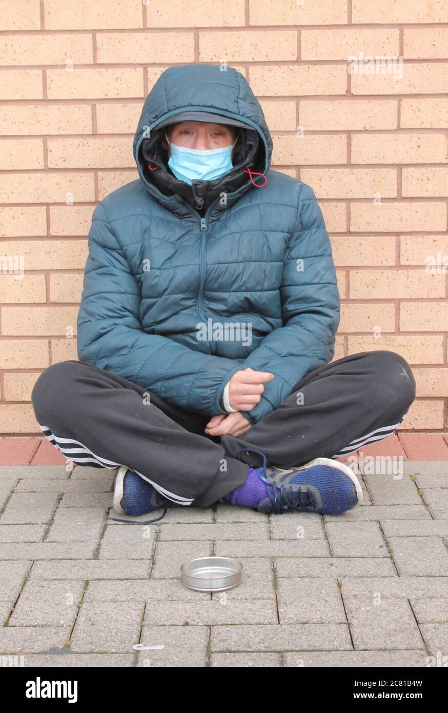 Homeless women with bruised eye, wearing a blue mask sitting on the floor with legs crossed begging for change Stock Photo