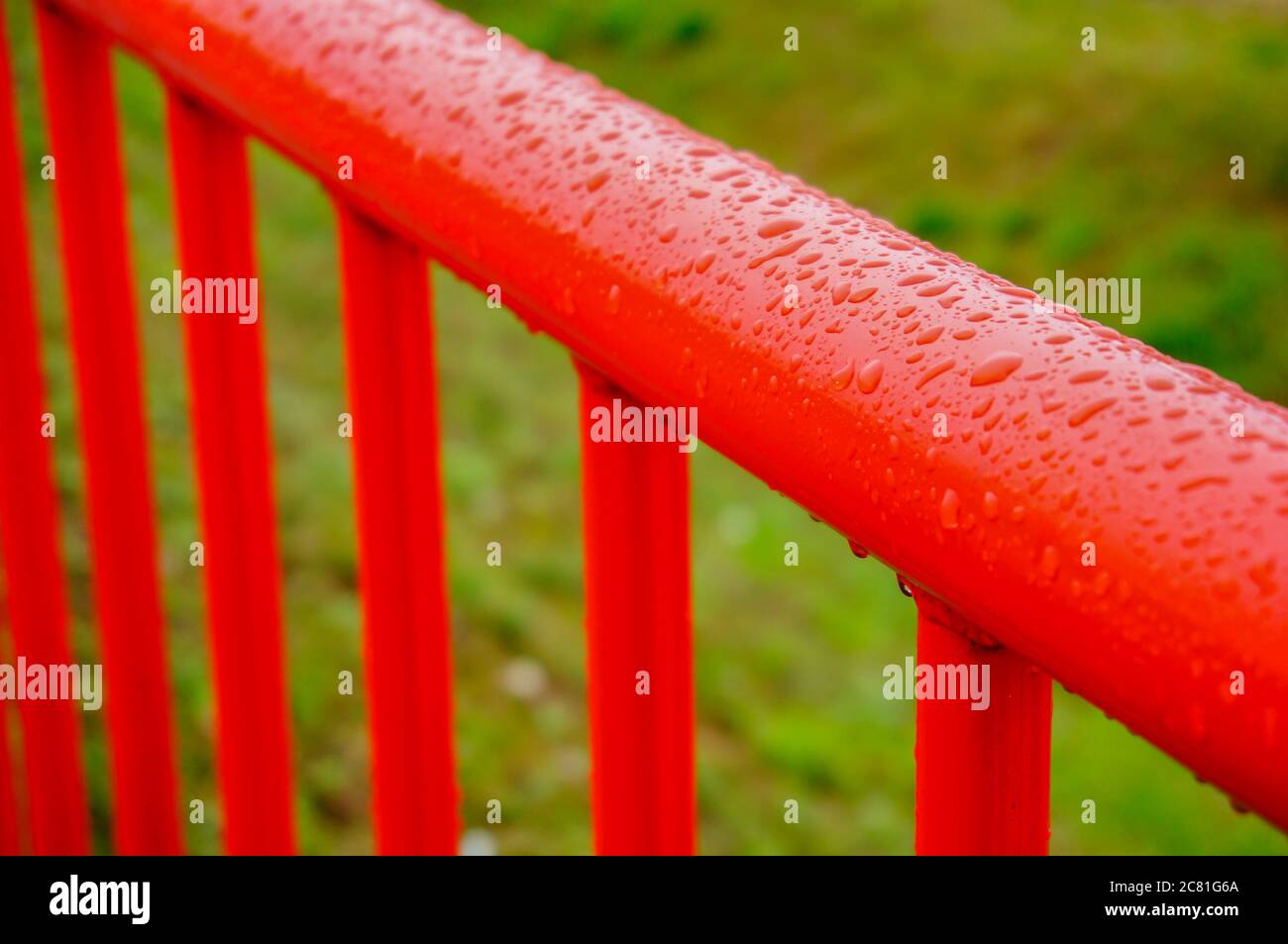 Closeup shot of a red metallic fence with vertical bars next to a green field on a rainy day Stock Photo