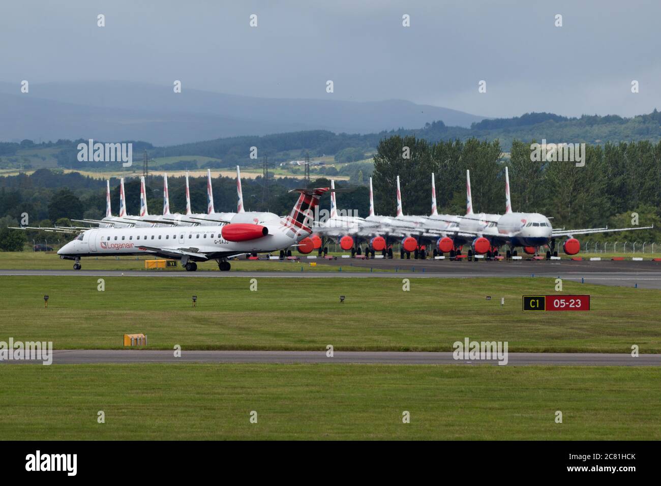 Glasgow, Scotland, UK. 20th July, 2020. Pictured: Loganair Embraer ERJ145 plane seen at Glasgow Airport with a large group of grounded British Airways (BA) airbus aircraft in the background. Loganair had taken over some of Flybe's slots after the collapse of Flybe, then the coronavirus lockdown hit in March, and since then Loganair have been increasing services but these are turbulent times for all airlines and the global aviation industry. Credit: Colin Fisher/Alamy Live News Stock Photo