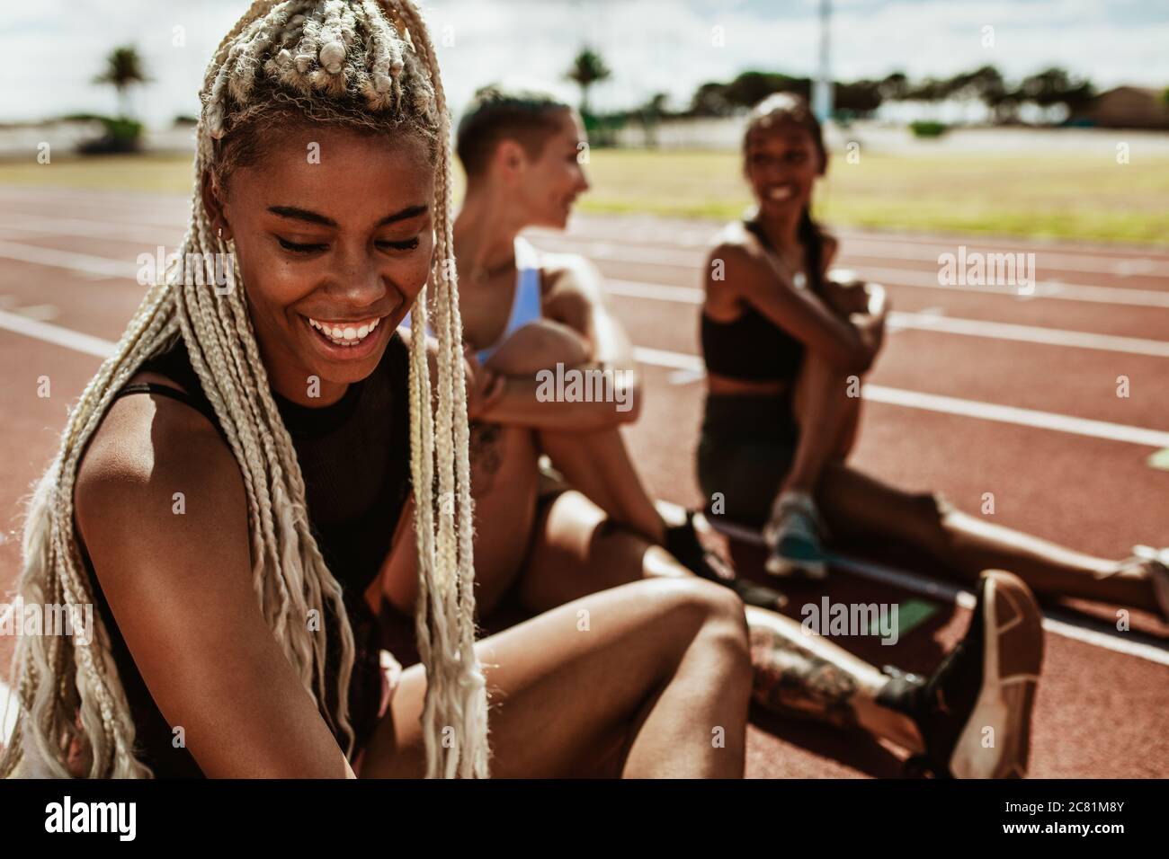 Smiling female athlete sitting on track with other runners stretching in background. Sportswoman relaxing and stretching after a practice run on the t Stock Photo