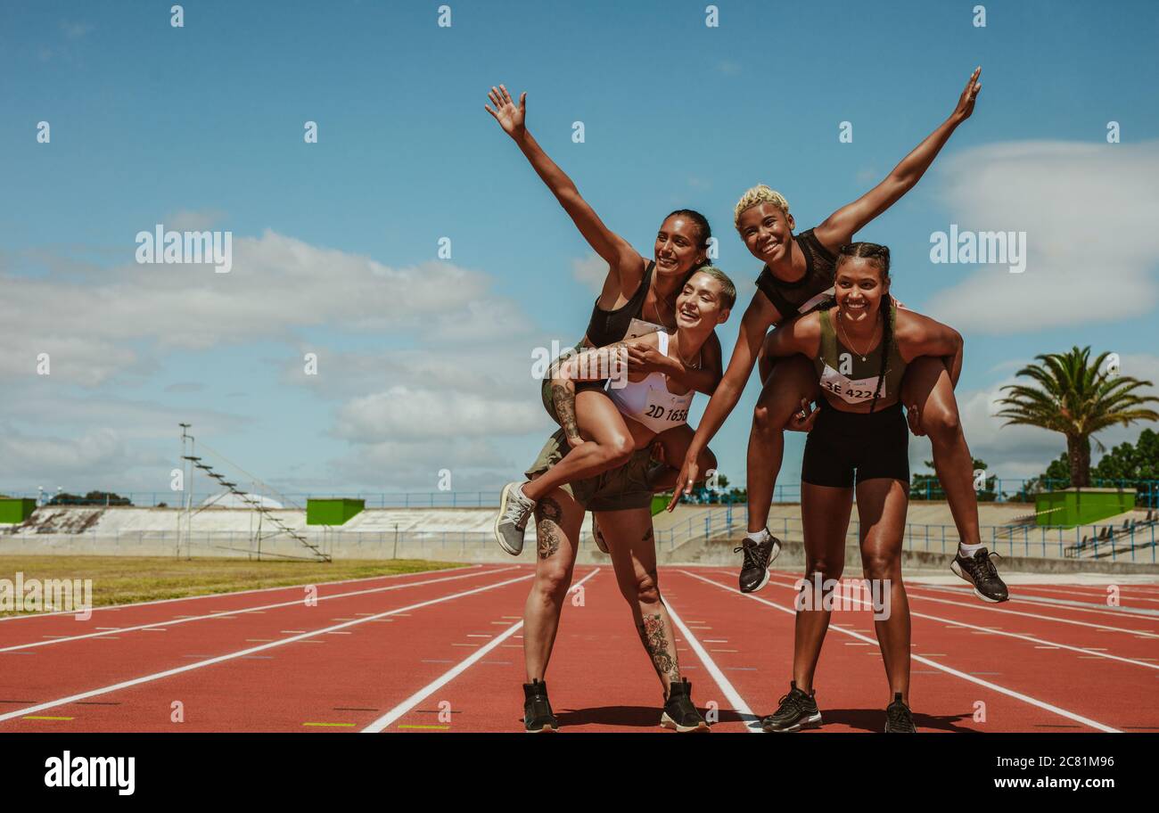 Female athletes carrying their teammate on back at stadium. Group of sportswoman enjoying on race track at stadium. Stock Photo