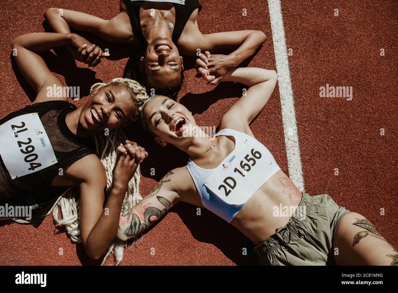 Top view of three female athletes lying together on running track holding hands and smiling while looking at camera. Successful team of runners enjoyi Stock Photo