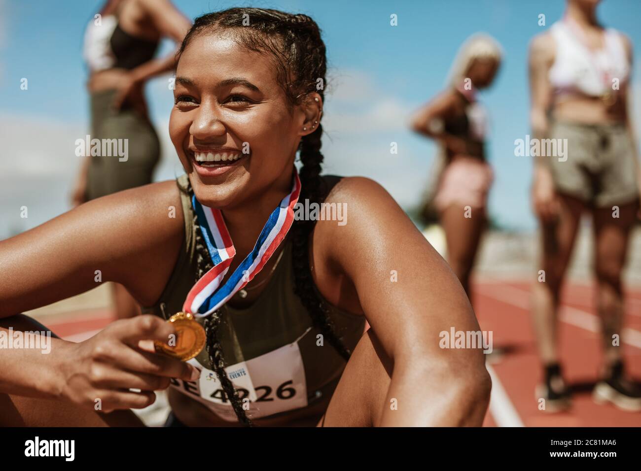 Female runner with a gold medal sitting on track. Running race winner sitting on track with athletes in background. Stock Photo