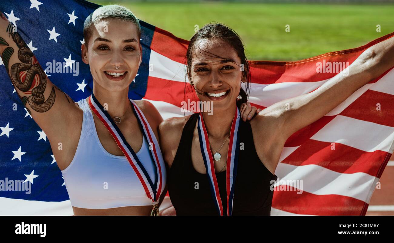 Two US athletes with national flag. Women runner with medals standing on running track holding American national flag. Stock Photo