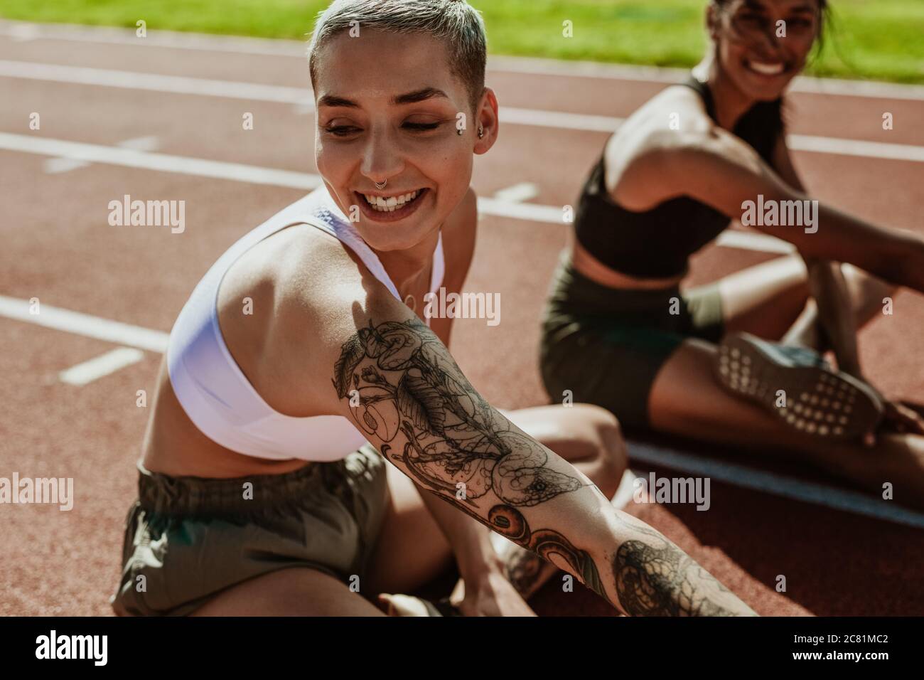 Runners stretching legs and smiling before a track event. Two young runners practicing at athletics stadium. Stock Photo