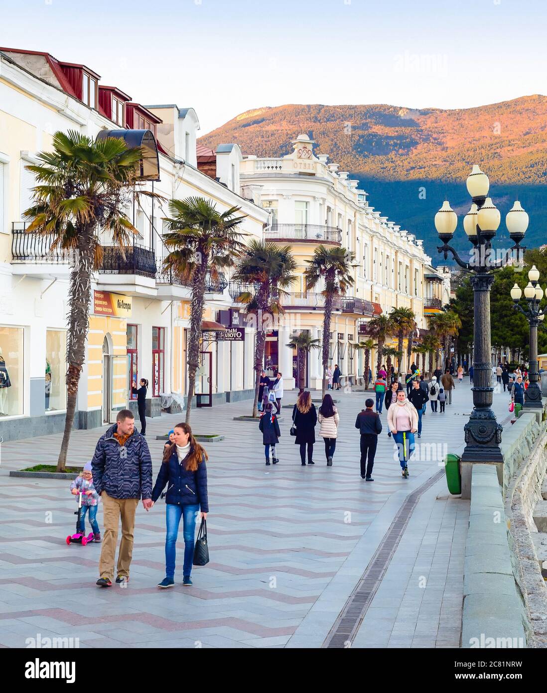 YALTA, CRIMEA - APRIL 3, 2018: People walking by resort city embankment, mountain in sunset light in background Stock Photo