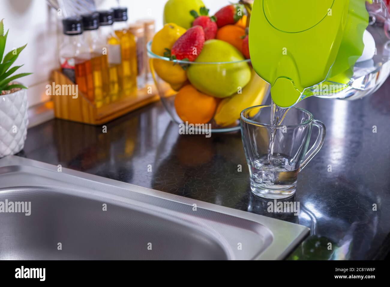 Pouring filtered water into glass from water filter jug in the kitchen. Purification and softening of drinking tap water. Closeup. Focus on glass. Stock Photo
