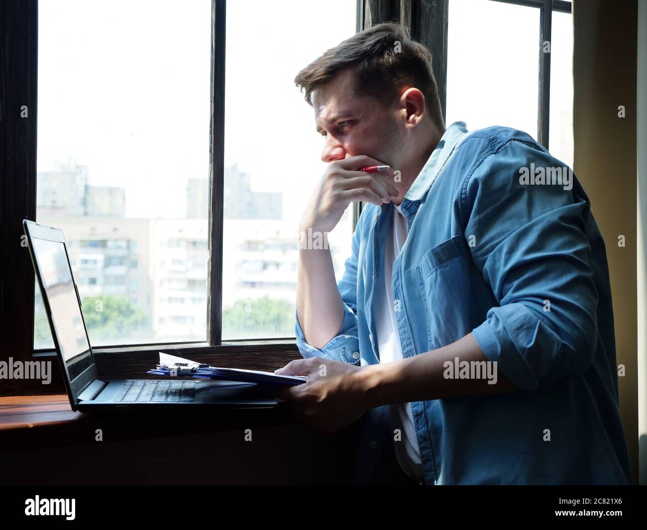 Young attractive man at the window working on laptop. Freelance and remote work. Stock Photo