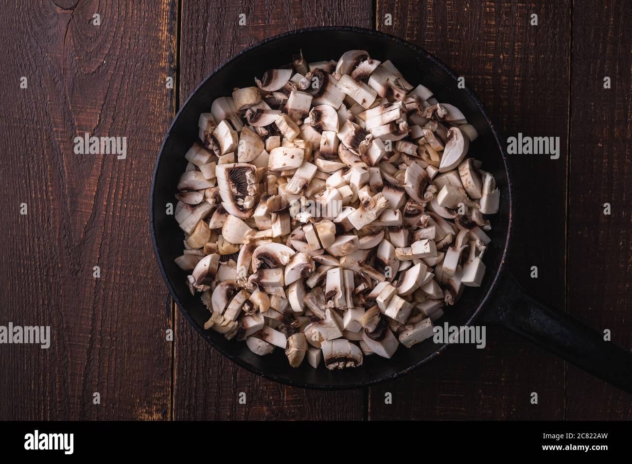 Tasty chopped champignon mushrooms in frying pan, uncooked food sliced, dark brown wooden background, top view Stock Photo