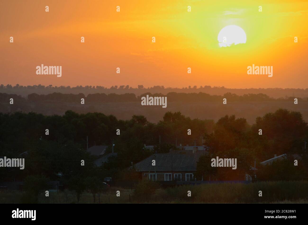 The white sun in the orange sunset of the evening haze above the village. Stock Photo
