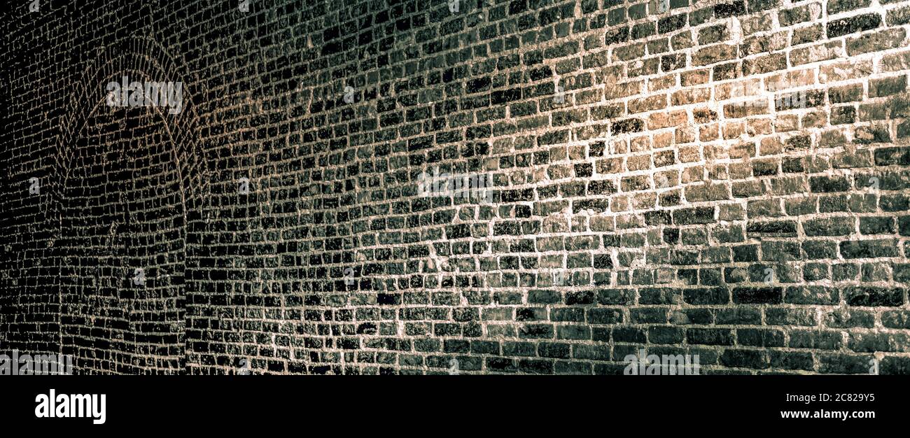 A panoramic format of a vintage brick wall with bricked up arched doorway, lit from dark to light in old basement in sepia tone Stock Photo