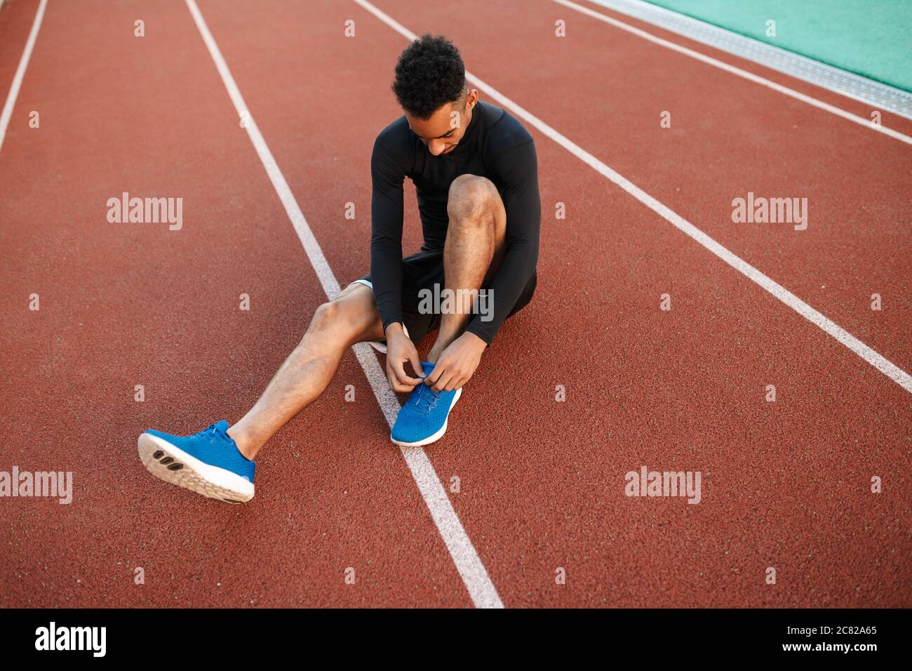 Young African American sporty man tying shoelaces on sneakers on racetrack at city stadium Stock Photo