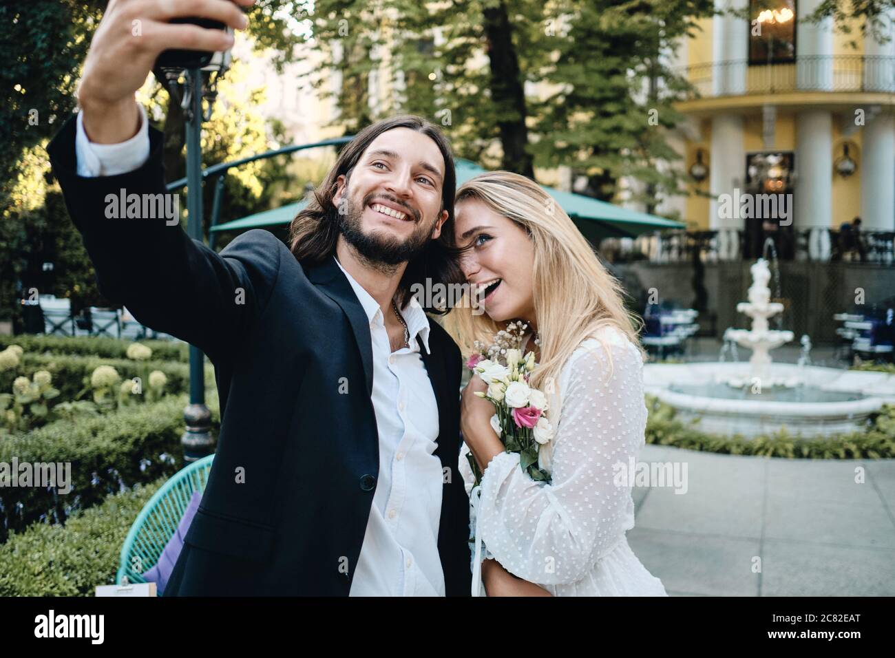 Beautiful romantic newlyweds happily taking selfie on cellphone in garden of restaurant Stock Photo