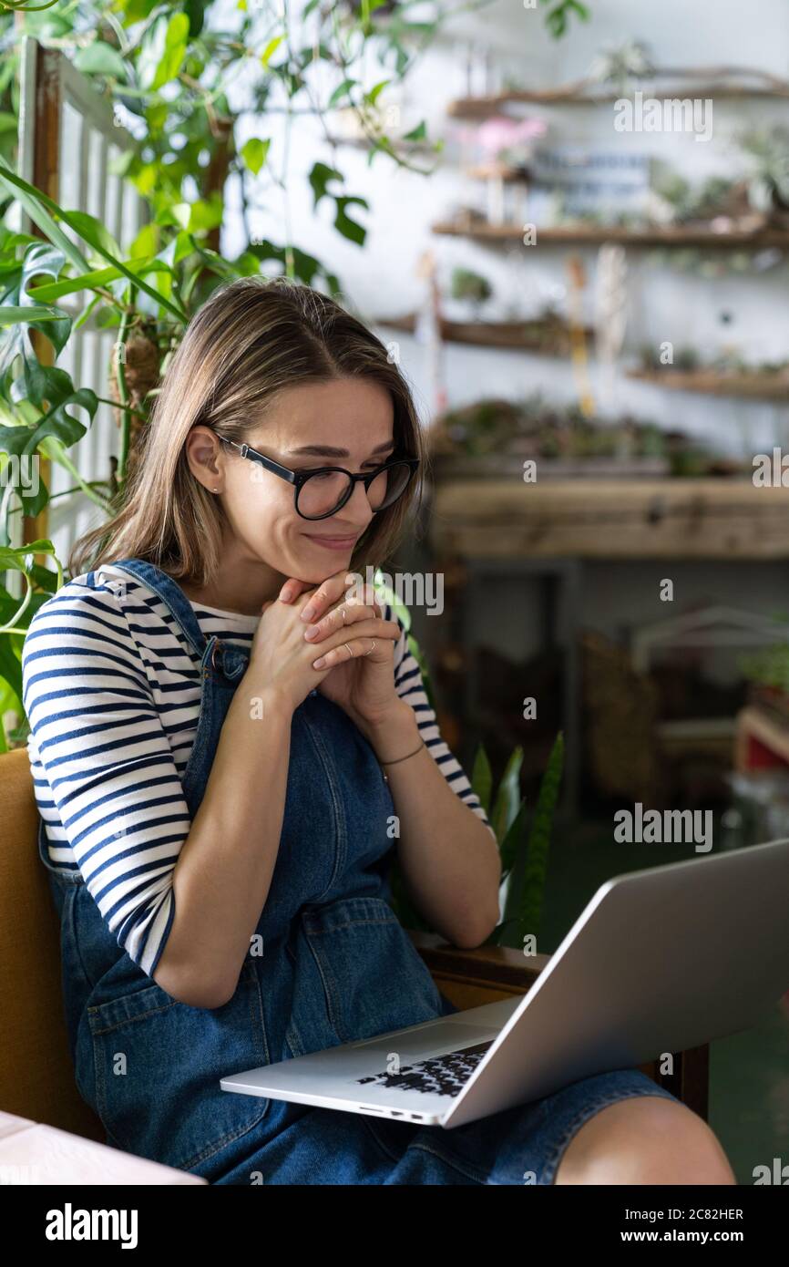Woman gardener in glasses wear blue jeans overalls, sitting on chair in greenhouse, using laptop after work, communicates on internet with customer su Stock Photo