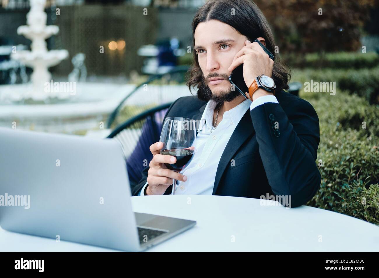Young serious confident bearded businessman with glass of wine talking on cellphone in restaurant outdoor Stock Photo