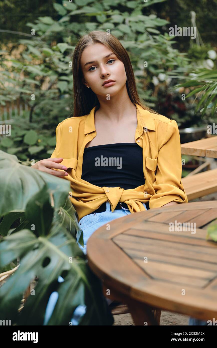 Young serious brown haired teenage girl in yellow shirt and top thoughtfully looking in camera while sitting at wooden table in city park Stock Photo