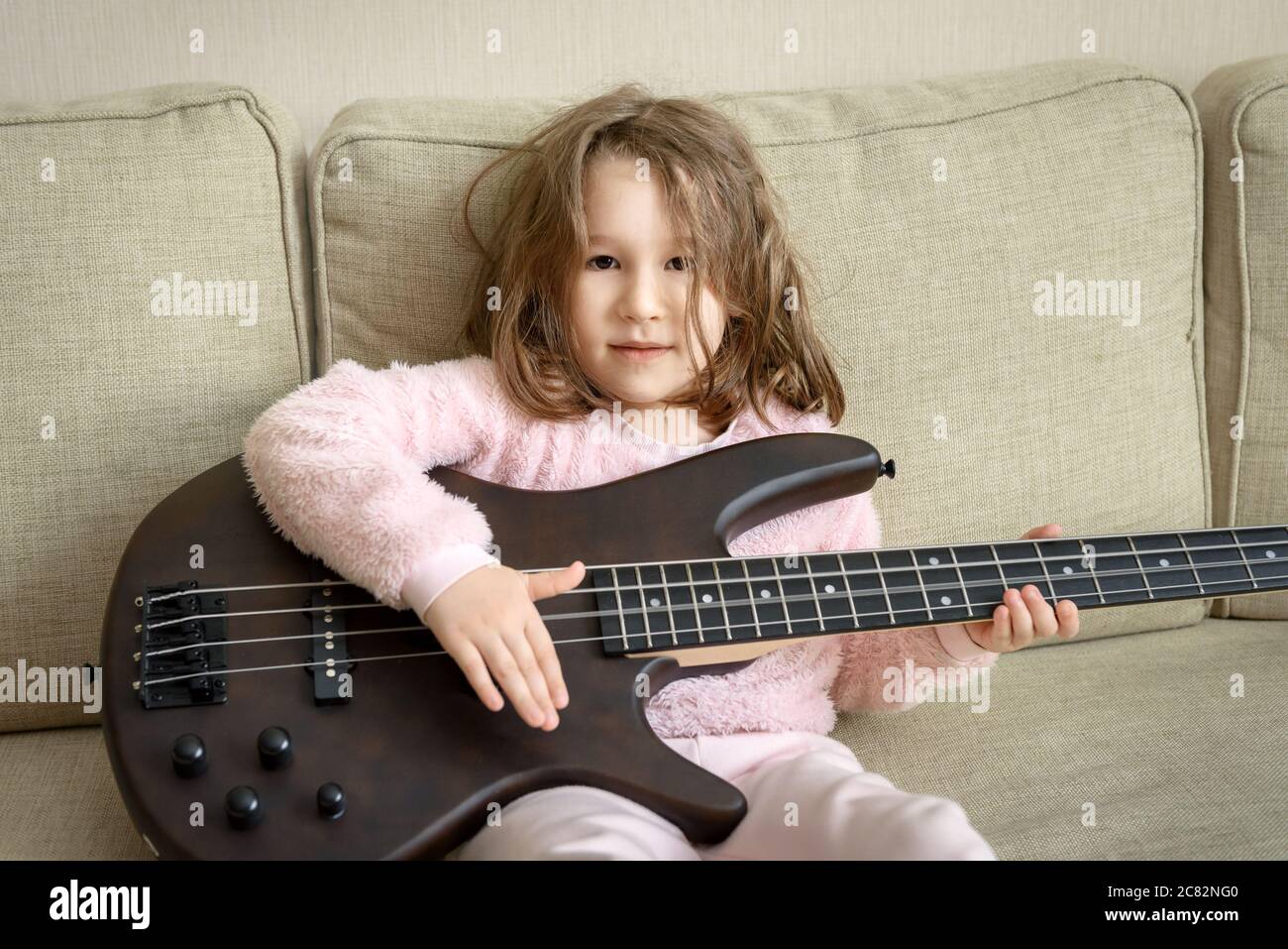 Child playing bass guitar at home, portrait of little baby girl sitting on couch with music instrument, happy cute kid hits the guitar strings, funny Stock Photo