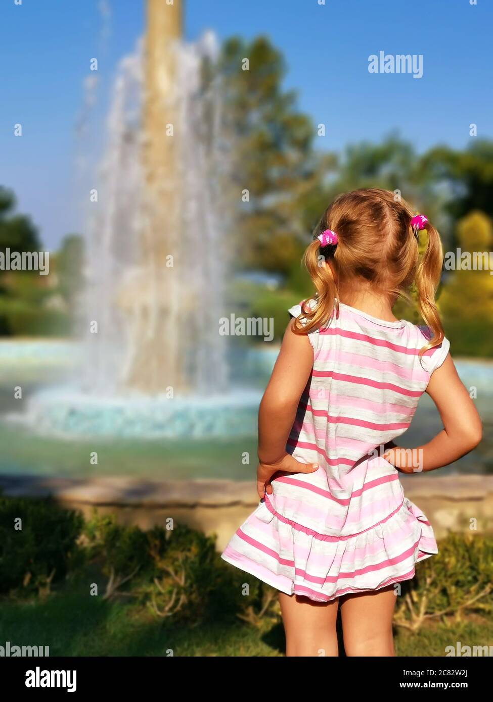 Preschooler little girl watching fountain rear view. Young female child enjoy being outside in summertime. Curiosity blonde looking at splashing dropl Stock Photo