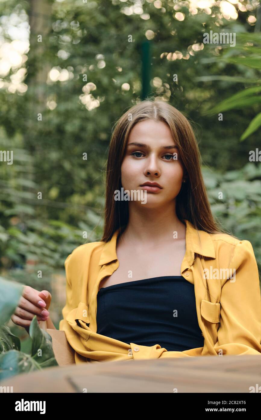 Young gorgeous brown haired woman in yellow shirt and top seriously looking in camera while sitting in city park Stock Photo