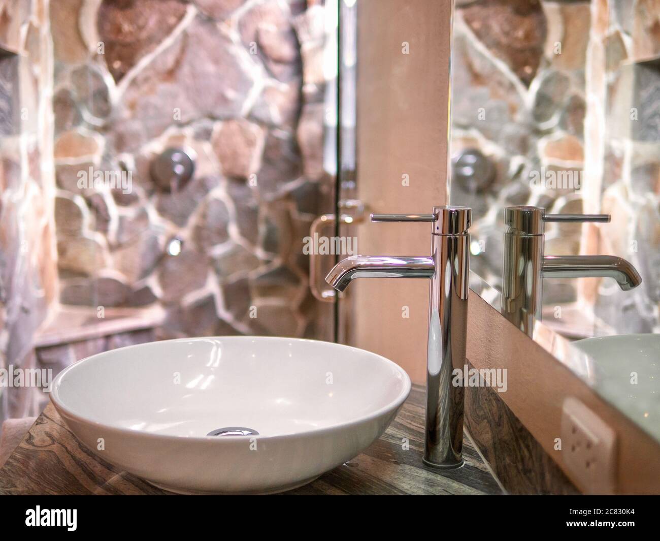 Close up view of a single white ceramic basin, polished chrome faucet, and mirror in a luxury hotel bathroom with a natural stone shower stall. Stock Photo