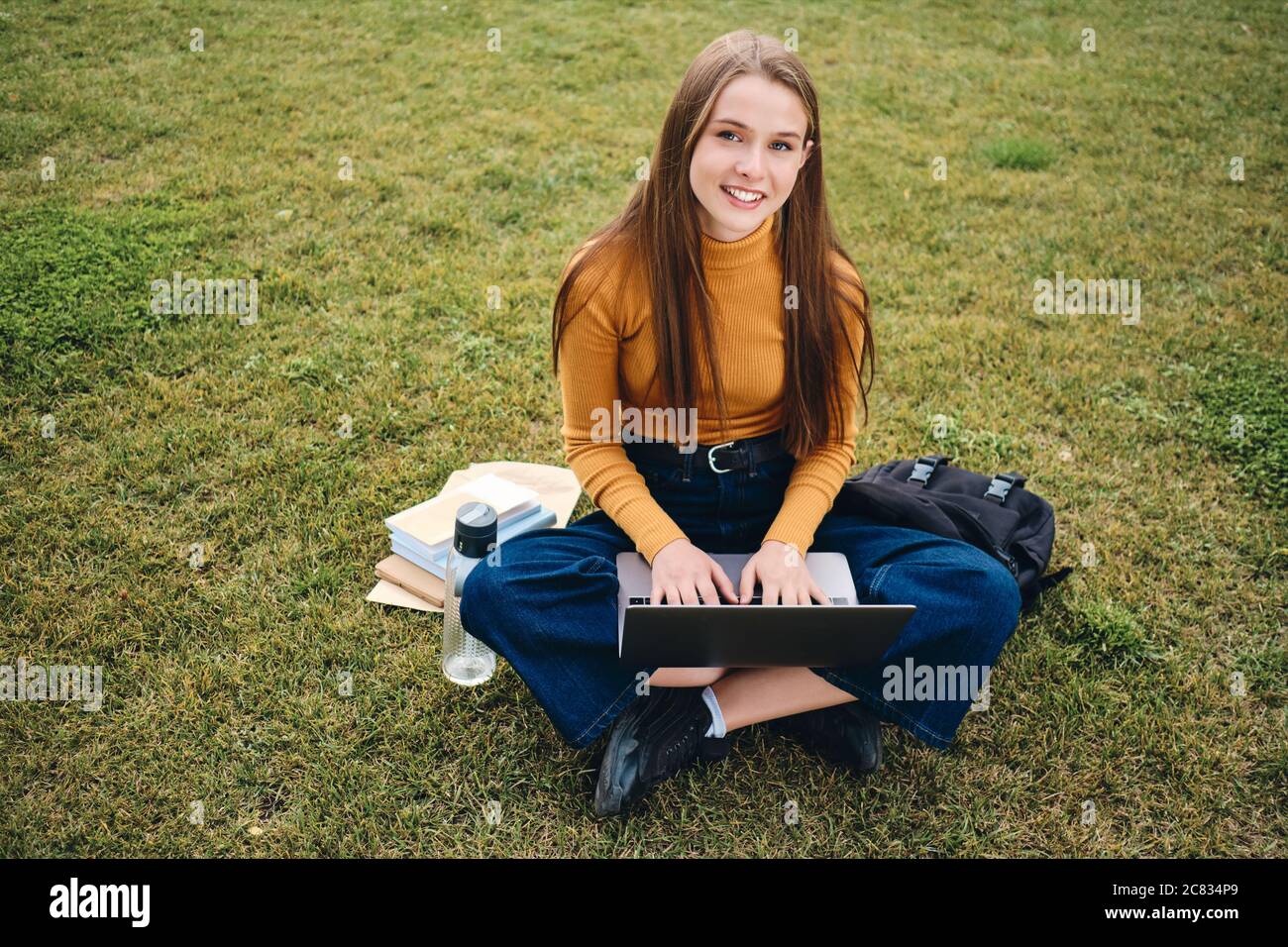 Pretty smiling casual student girl happily looking in camera studying with laptop on lawn in city park Stock Photo