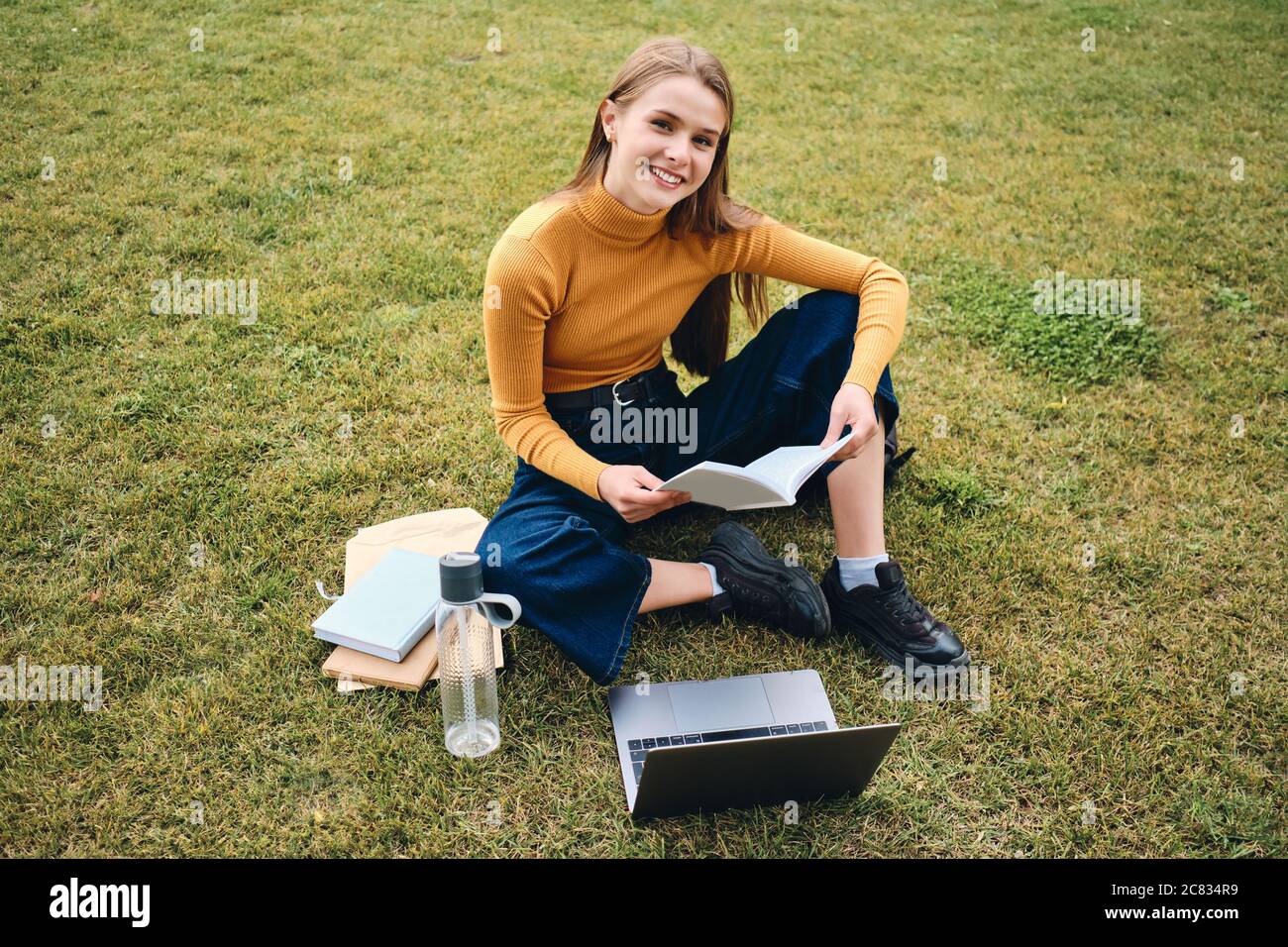 Attractive cheerful student girl happily looking in camera studying on lawn in city park Stock Photo