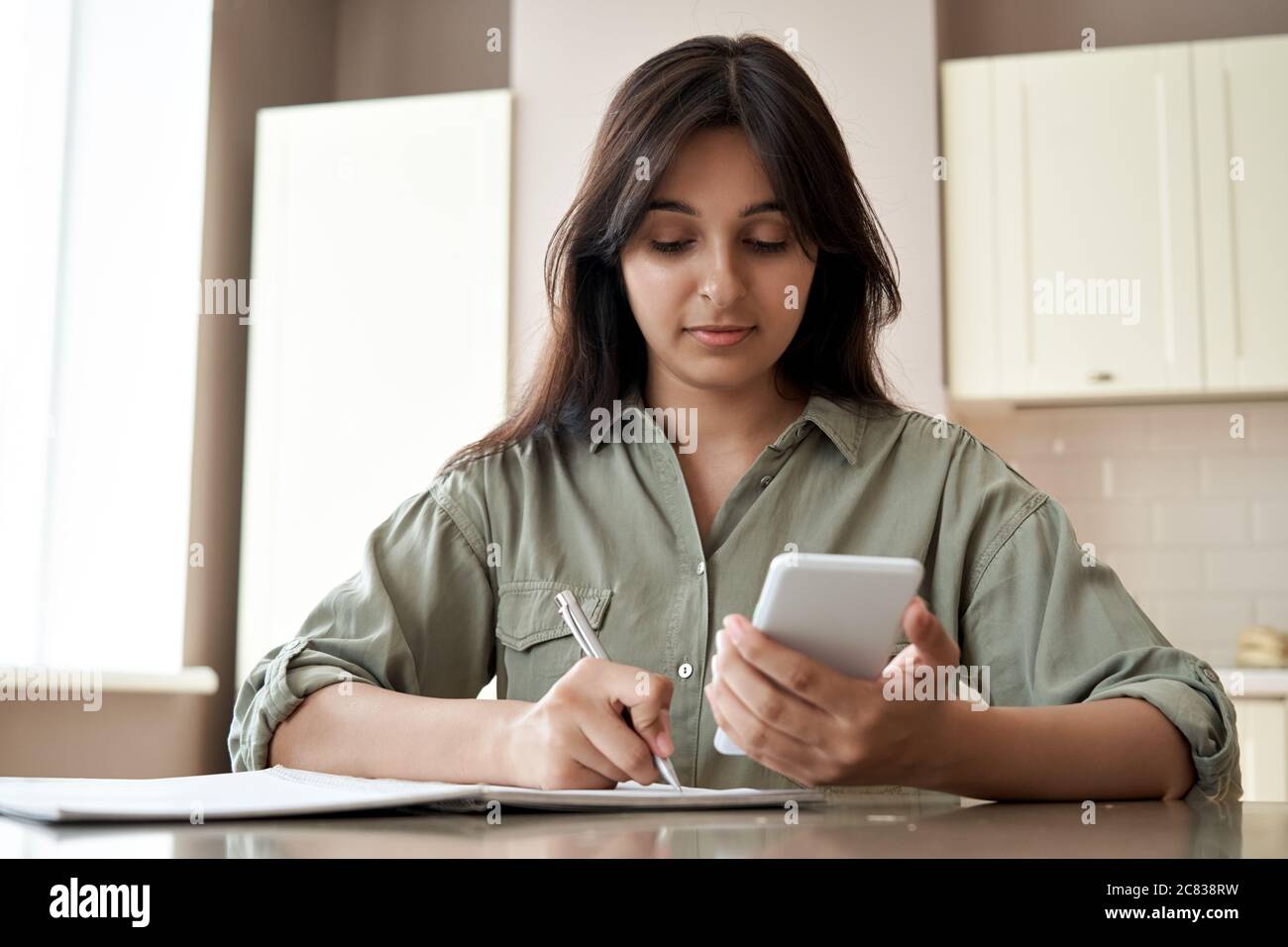 Indian girl holding phone online learn in mobile app write notes at home office. Stock Photo