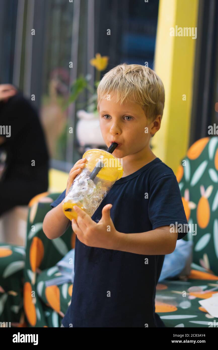 Front view of happy thirsty blond kid boy drinking lemonade, fruit tea, kombucha ice tea with peach and lemon, wellness orange drinks from plastic cup Stock Photo