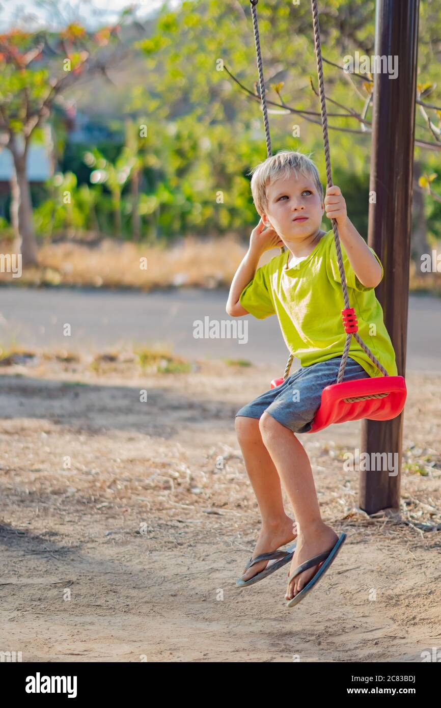 Sad lonely boy sit on swing look far away, wait for friends or while parents are busy, thinking about important things for him. Summer, childhood Stock Photo