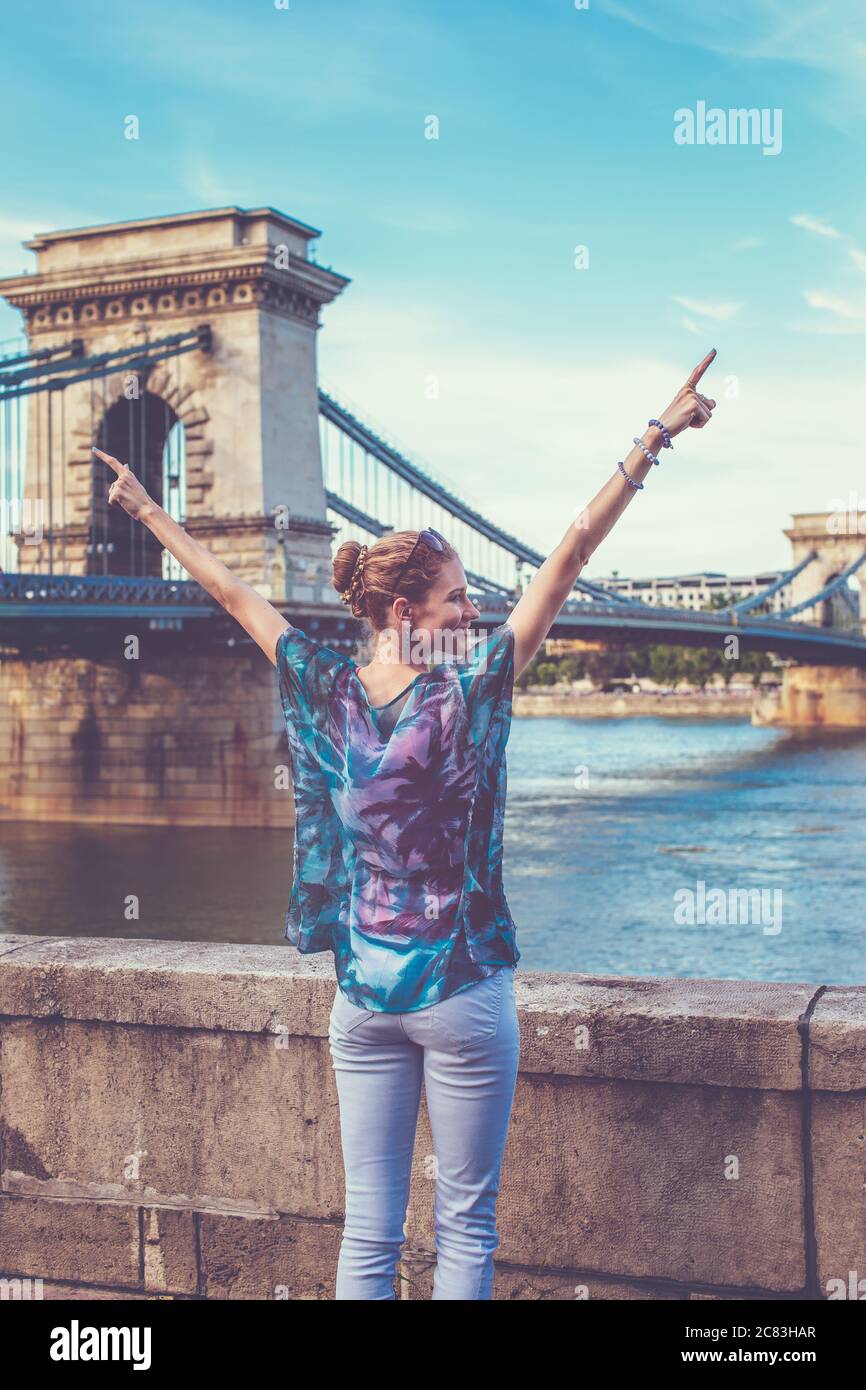 Young redhead woman arms raised and looking away at Chain Bridge, Budapest, Hungary Stock Photo