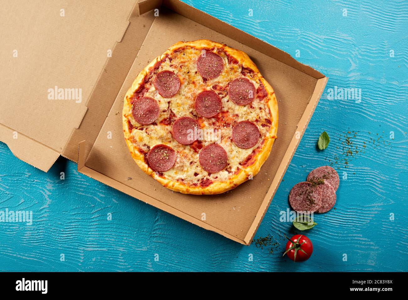 Oven-fired traditional Italian pepperoni or salami pizza in a cardboard box ready for takeaway with fresh ingredients alongside on blue wood in an ove Stock Photo